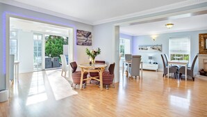 A spacious living area with a wooden floor, a dining table with chairs, and a view of greenery outside. - The Coast House B&B (Bognor Regis)
