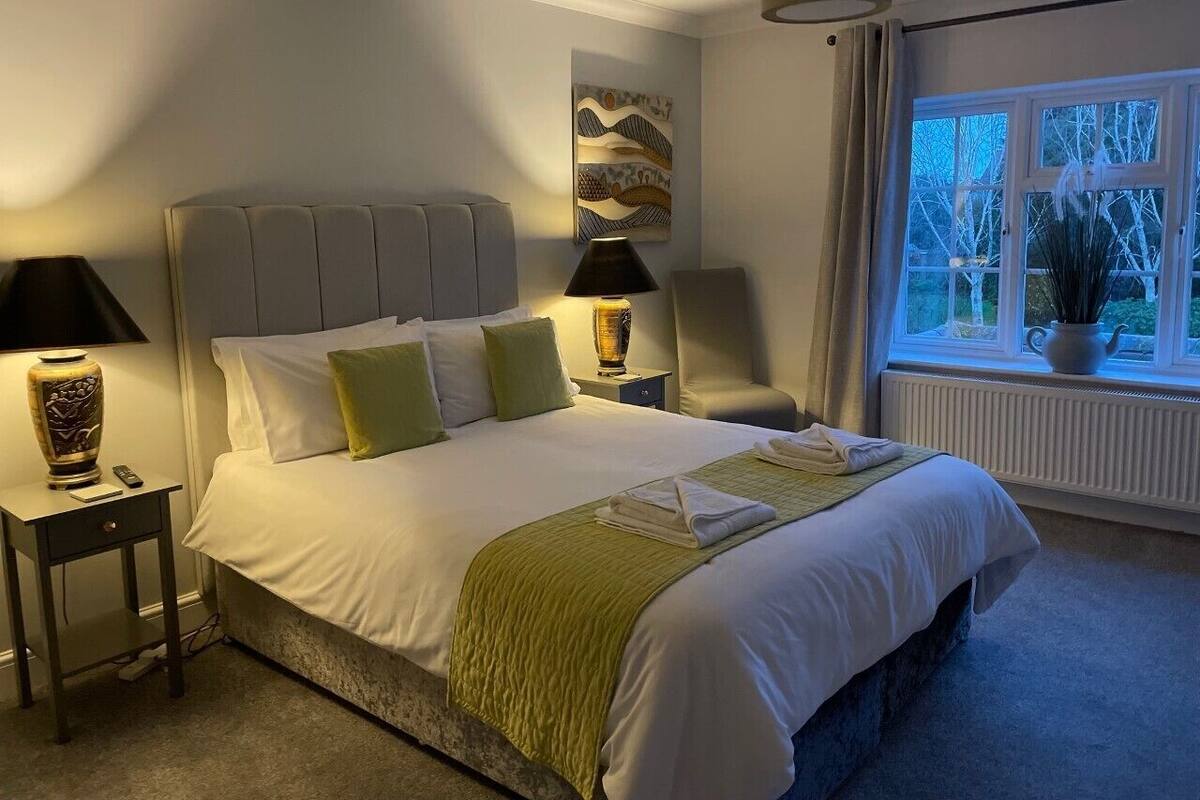Una cama bien hecha con una colcha verde, dos lámparas de noche y una ventana con vistas a árboles.