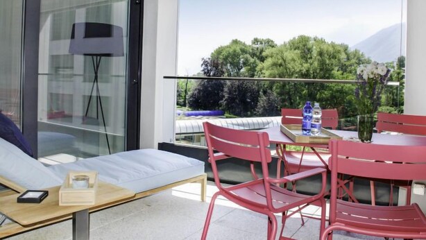 A balcony with a sofa, a dining table, and chairs. There is a view of trees and mountains in the background. - LaVille B-1-2 by Interhome (Locarno)