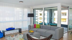 A modern living room with a grey sofa, a small table with fruit, a floor lamp, and a view of a building through glass doors. - LaVille B-1-2 by Interhome (Locarno)
