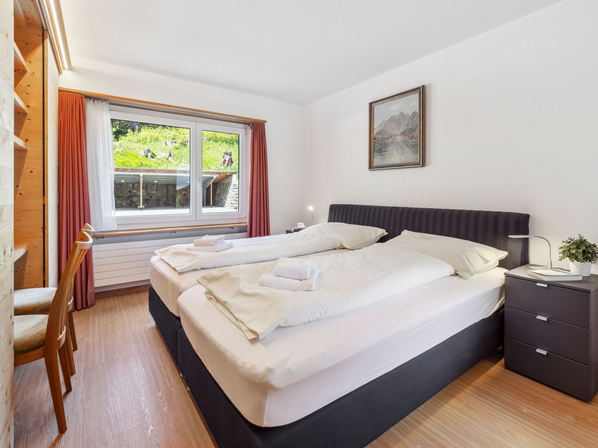 Uma cama de casal com roupa de cama branca, uma mesa de cabeceira com abajur e uma janela com cortinas.