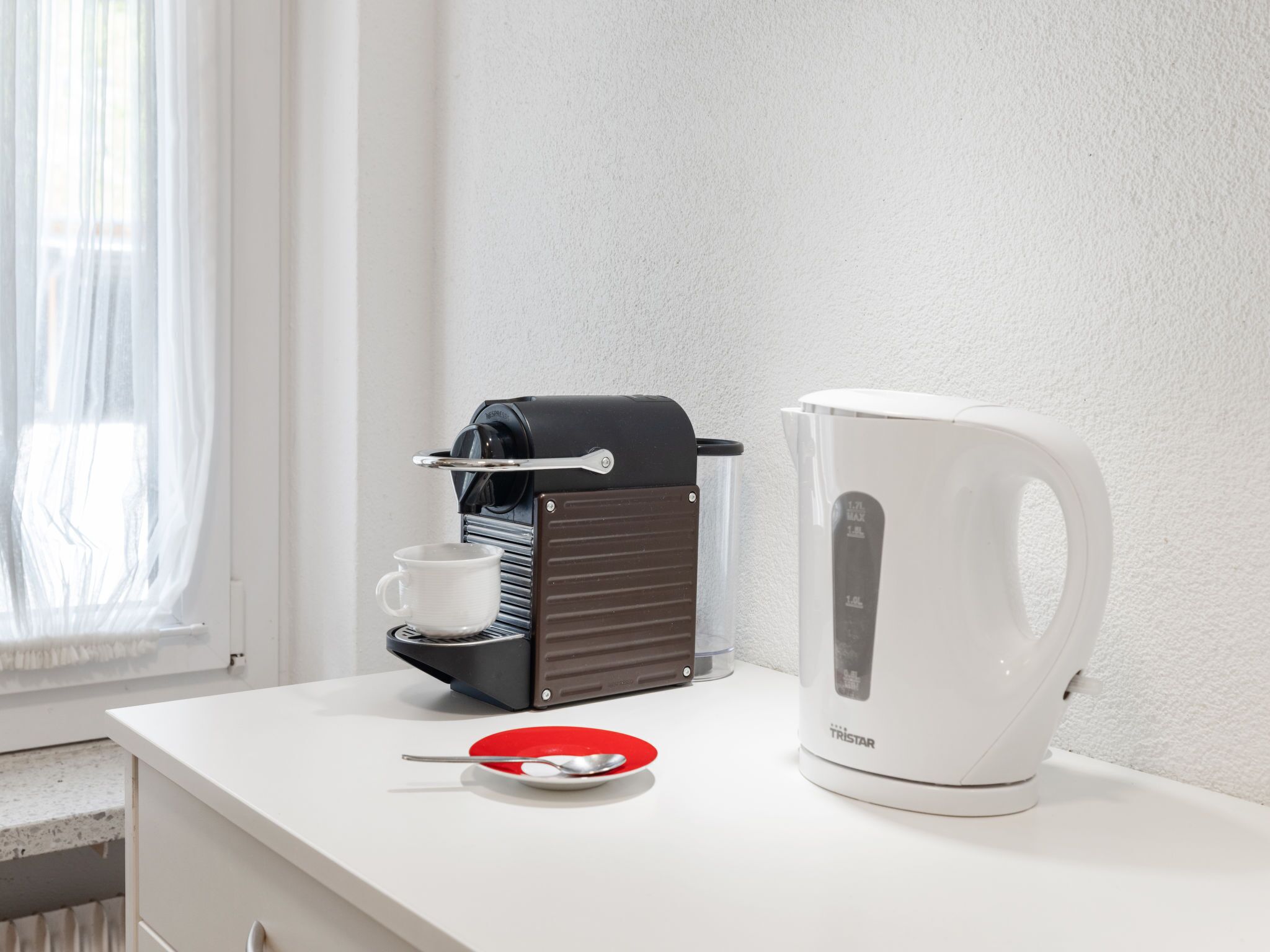 A coffee maker, a white electric kettle, and a red plate with a spoon on a white countertop near a window.