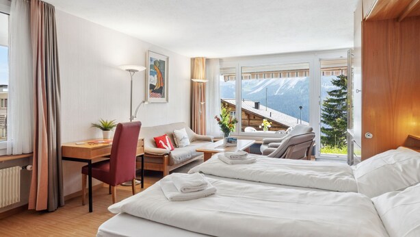A hotel room with a bed, a desk, a chair, and a view of mountains through large windows.