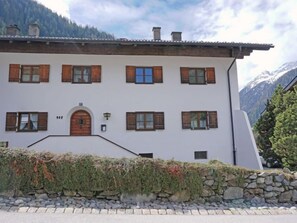 Un bâtiment blanc avec des volets en bois, un mur de pierre et une chaîne de montagnes en arrière-plan.