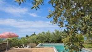 Un espace aménagé au bord de la piscine, avec deux chaises longues, un parasol rouge et blanc à rayures, et une vue sur la piscine et la végétation environnante.