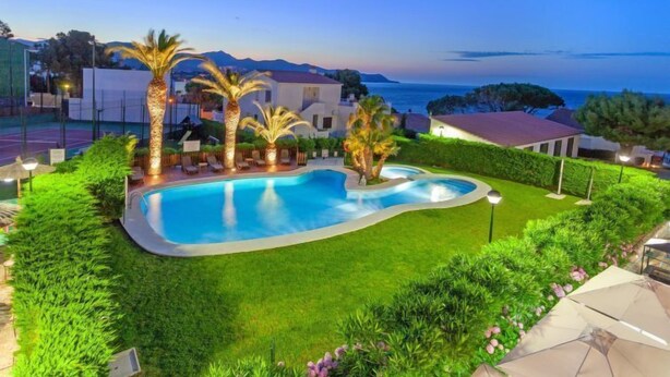 A swimming pool surrounded by palm trees and greenery, with a view of the sea and mountains in the background. - Palmiers I by Interhome (Llançà)