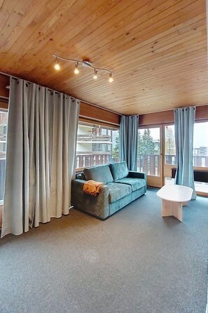 Une pièce avec un plafond en bois, un canapé gris, une petite table blanche et une vue dégagée sur l’extérieur grâce à de grandes fenêtres.