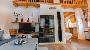 Un salón amplio con techo alto, vigas de madera, balcón, televisor de pantalla plana, chimenea y cocina al fondo.