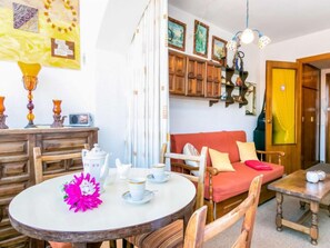 A cozy living room with a round dining table, wooden chairs, a red sofa, and a kitchen area with wooden cabinets and a hanging light fixture. - Roses Holiday Center by Interhome (Roses)