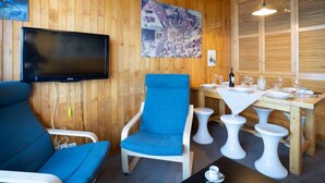 Un intérieur en bois avec un téléviseur, des chaises bleues, une table à manger agrémentée de tabourets blancs et un tableau accroché au mur.