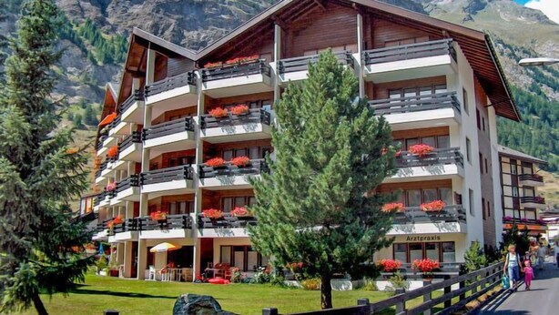 Un hotel de diverses plantes amb balcons, envoltat de muntanyes i vegetació.