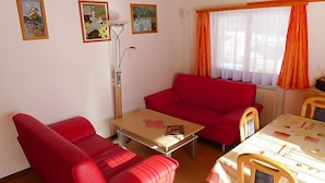 A room with a red sofa, a small table, and a window with orange curtains. There are three framed pictures on the wall. - Christl by Interhome (Stalden)