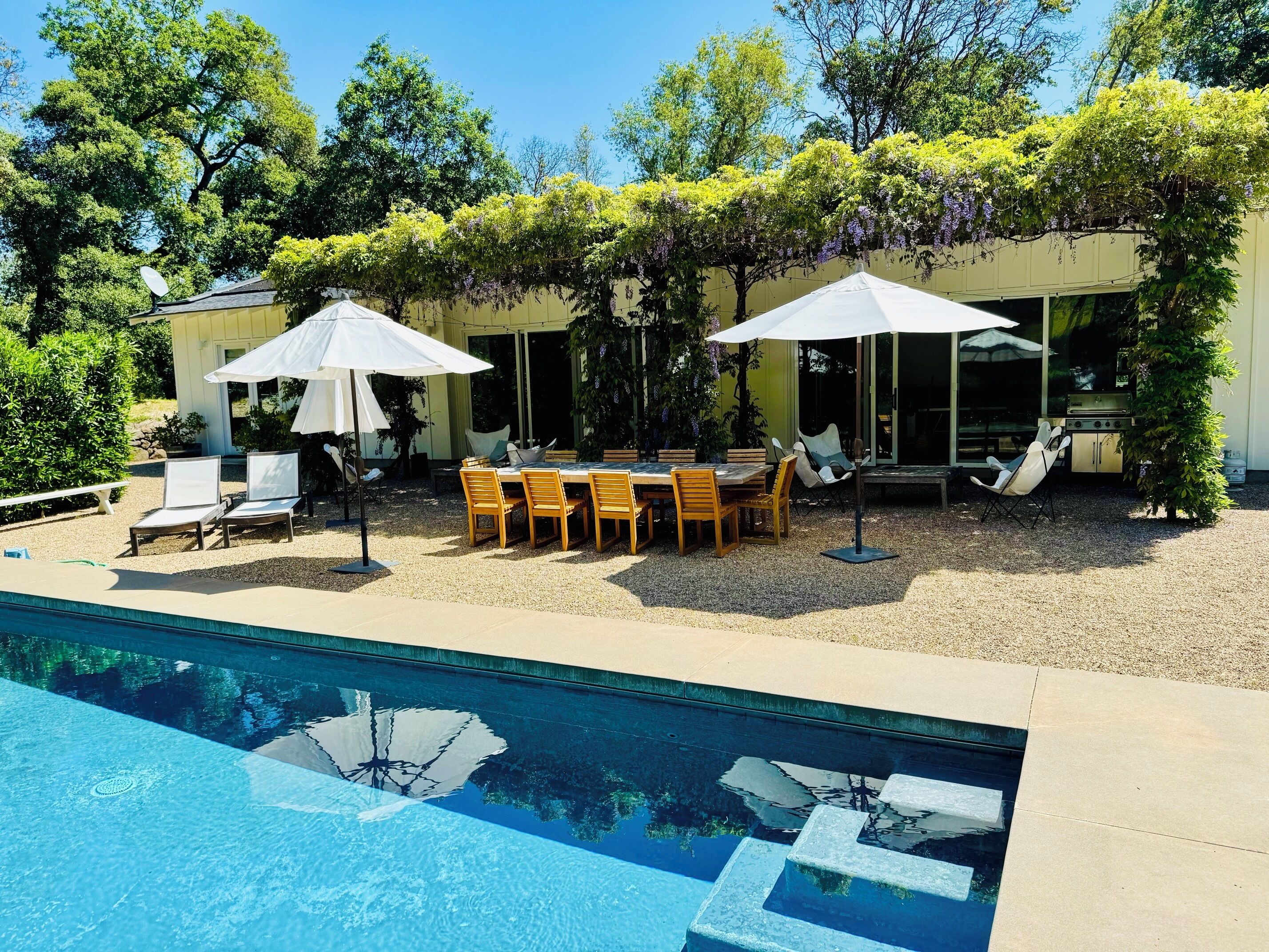 A poolside area with a table and chairs, umbrellas, and lounge chairs.