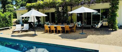 A poolside area with a table and chairs, umbrellas, and lounge chairs.