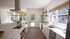 Une cuisine moderne avec des meubles blancs, un grand îlot central doté d’un évier et une vue sur l’extérieur grâce à de grandes fenêtres.