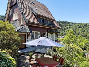 Une maison à deux étages avec un toit en pente, une terrasse avec une table et des chaises, et une vue sur une colline boisée.