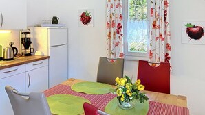 A small, modern kitchen with a refrigerator, coffee maker, and kettle. There is a dining table with a vase of flowers and a red chair.