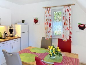 Une petite cuisine moderne équipée d’un réfrigérateur, d’une machine à café et d’une bouilloire. On y trouve une table à manger ornée d’un vase de fleurs et une chaise rouge.