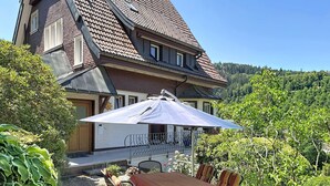 A house with a tiled roof, a balcony with a table and chairs, and a garden with various plants.