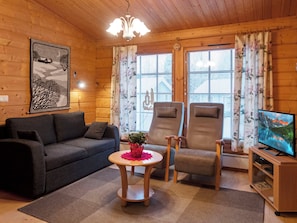 Interior acogedor de una cabaña con sofá, dos sillones, una mesita con una planta, televisor y techo de madera.