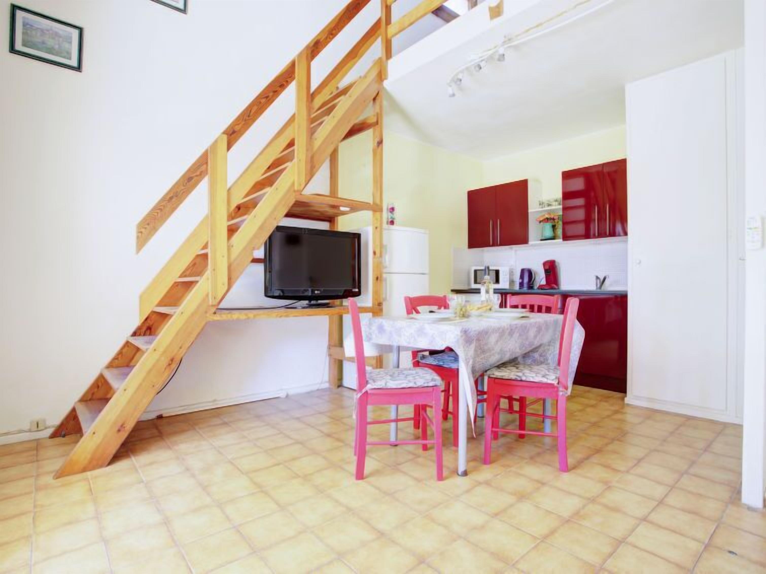 A compact living space with a wooden staircase, a dining area with pink chairs, and a kitchen with red cabinets.