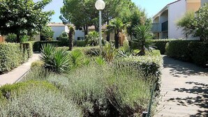 A pathway flanked by neatly trimmed hedges and various plants, leading to a building with a white facade and a spherical light fixture.