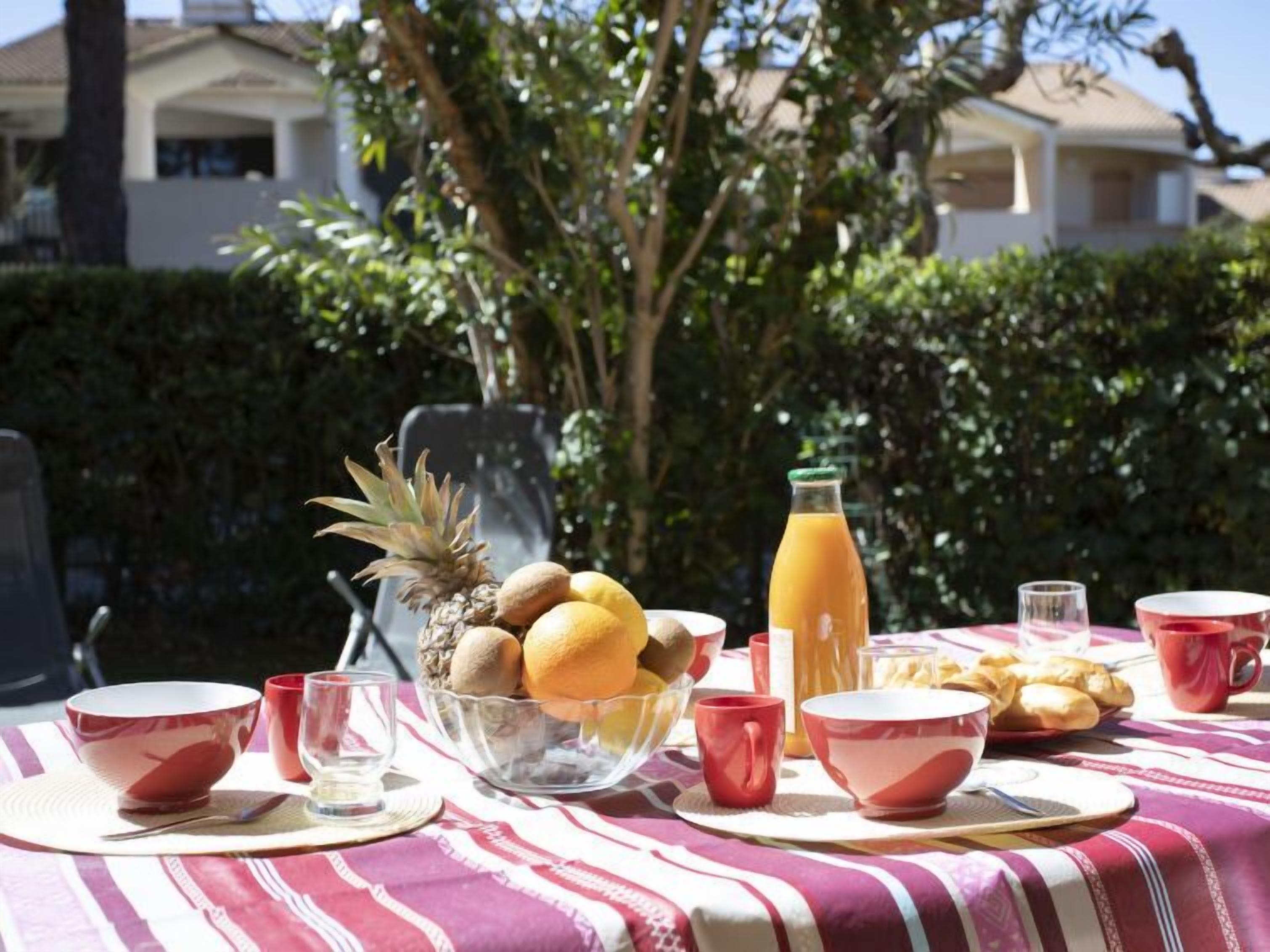 A breakfast table set outdoors with fruits, a bottle of orange juice, and red cups.
