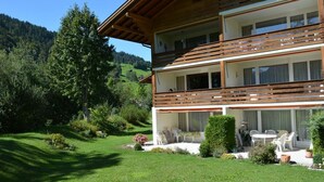 Une maison en bois à deux étages, dotée d’un balcon, entourée d’une végétation luxuriante et avec une chaîne de montagnes en toile de fond.