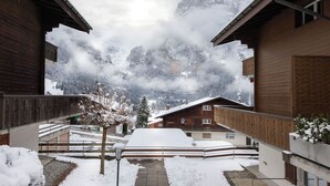 Ein verschneiter Innenhof mit Holzbauten, ein schneebedeckter Baum und ein Berg im Hintergrund.