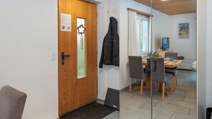 Ein modernes Zimmer mit Holztür, kleinem Fenster, Essbereich mit Tisch und Stühlen sowie einem Spiegel, der den Raum reflektiert.