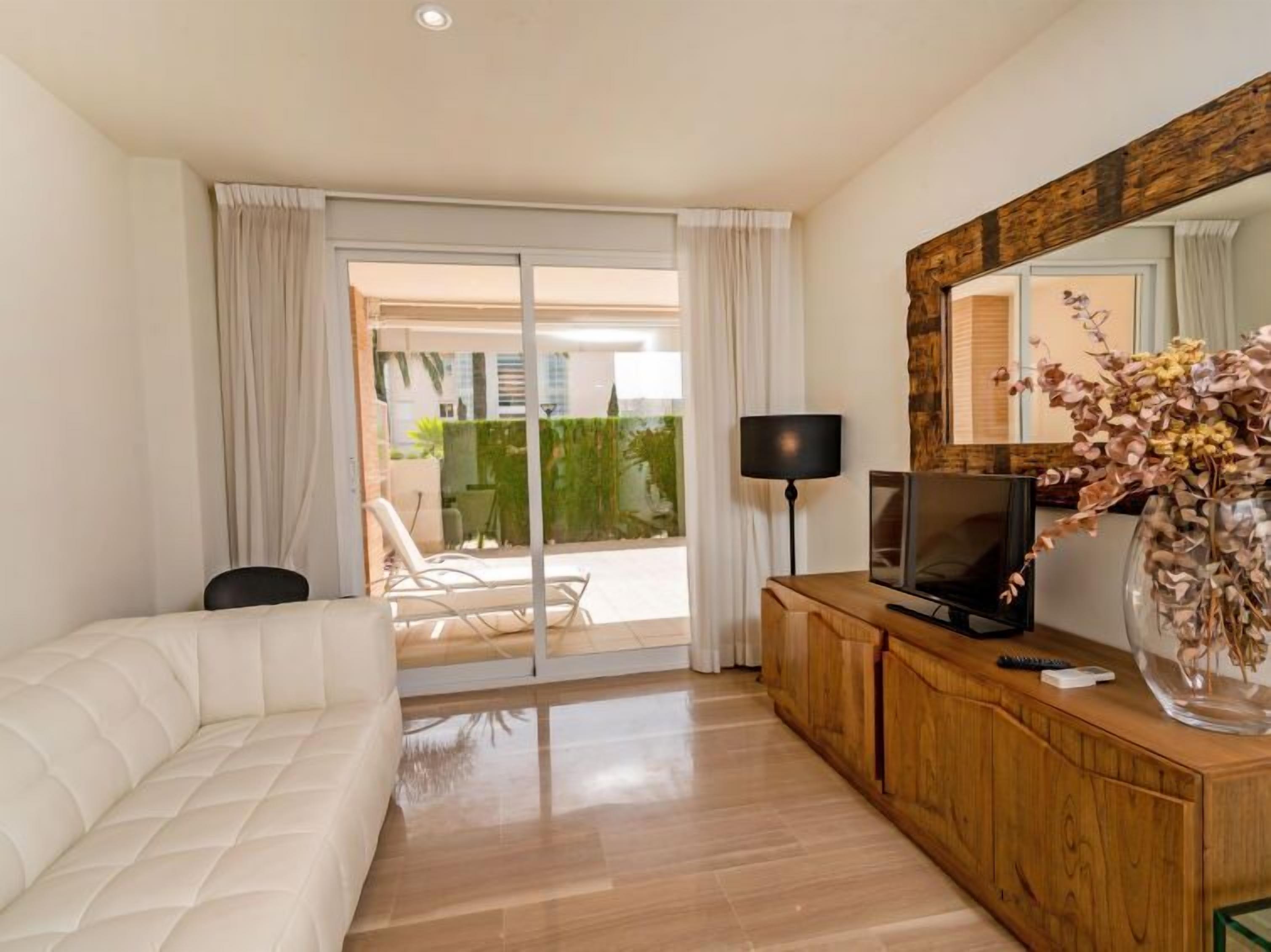 A modern hotel room with a white sofa, a wooden TV stand, a flat-screen TV, a lamp, and a large mirror.