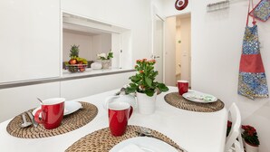 En stilren, modern matplats med vita väggar, ett vitt bord dukat med röda muggar, tallrikar och en vit kanna. Det finns vävda bordstabletter, en krukväxt och en röd handduk som hänger på väggen.