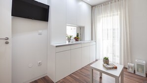 Ett modernt, minimalistiskt rum med en inbyggd TV, ett litet bord med dekorativa föremål och ett fönster med tunna, genomskinliga gardiner.