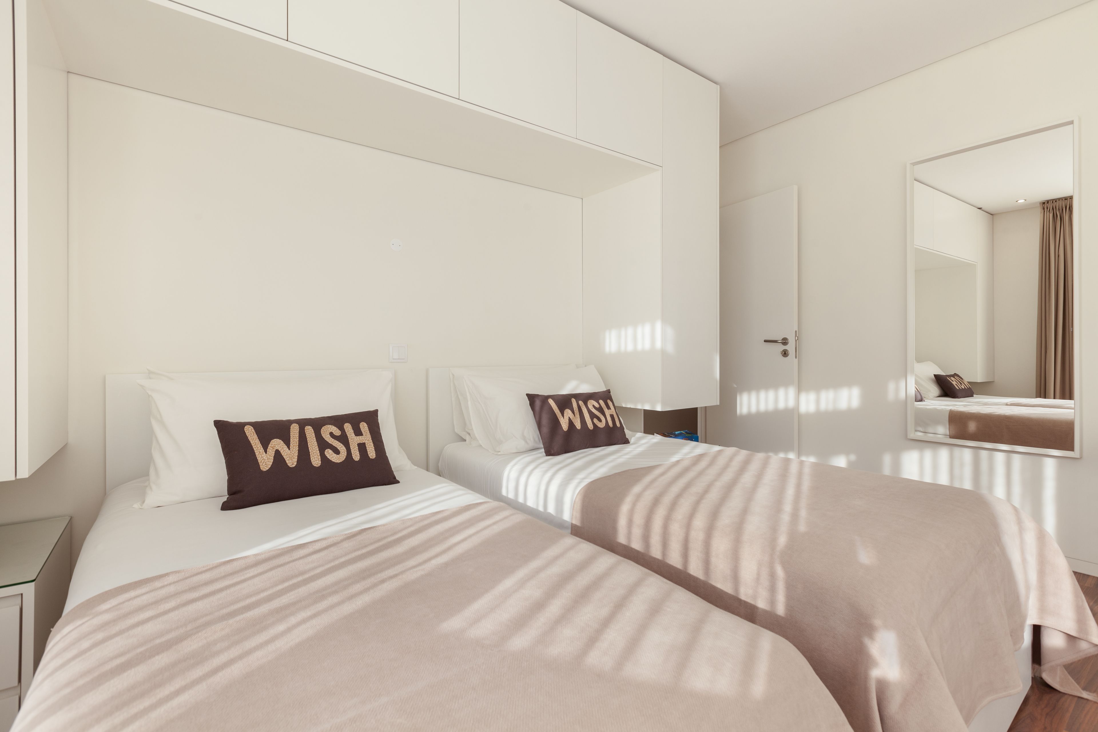 Quarto de hotel com duas camas, cada uma com uma almofada que diz 'WISH', um espelho grande e um guarda-roupa.