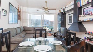 A modern living room with a glass dining table set for four, a sofa, a ceiling fan, and a bookshelf with books and decorative items. - Windsor by Interhome (Blanes)