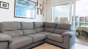 A grey sectional sofa in a modern living room with a large window, a framed painting, and a small table with a chair. - Windsor by Interhome (Blanes)