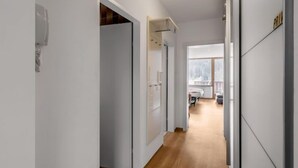 Een moderne hal met houten vloer, witte muren en zicht op een kamer met een bed en een raam.