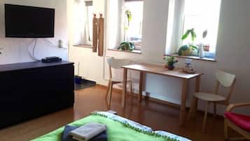 A bedroom with a bed, a desk, a chair, a TV, and a window with plants.
