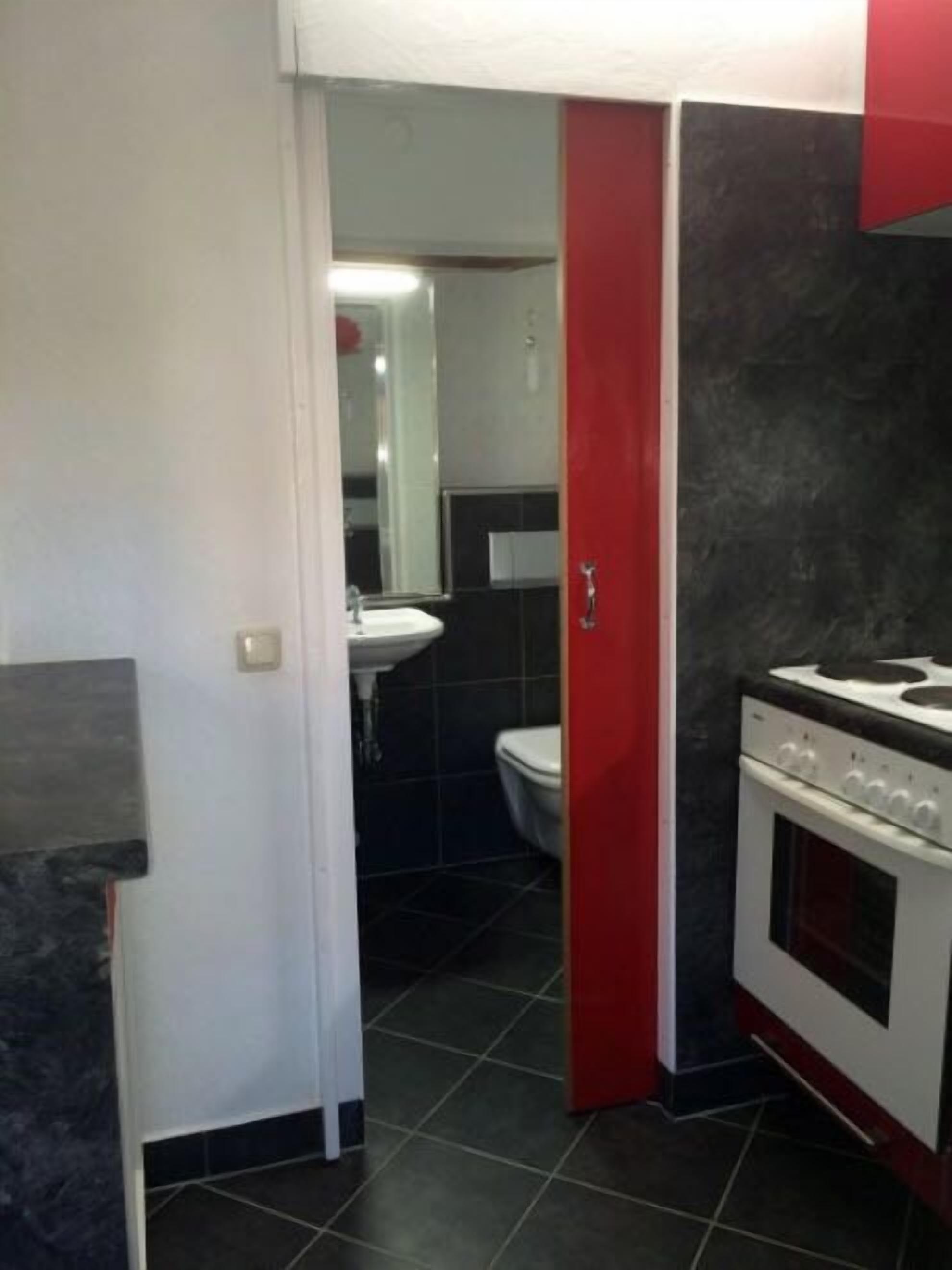 A compact kitchen with a stove, sink, and a red door leading to a bathroom.