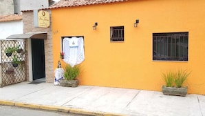 A building with a yellow facade, two small windows, and a door. There are potted plants on the sidewalk and a sign above the door. - Pousada Pai Nosso (Aparecida)