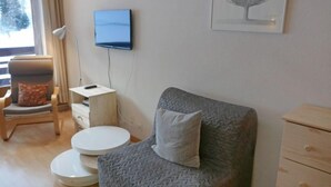 A modern living room with a grey armchair, a small white side table, a round white table, a wooden chair with a red cushion, a mounted flat-screen TV, and a framed artwork on the wall.