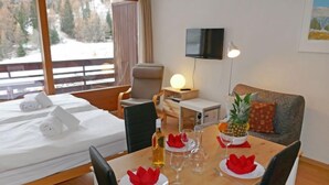 A hotel room with a bed, a wooden table set for dining, a television, and a window with a view of trees and snow.