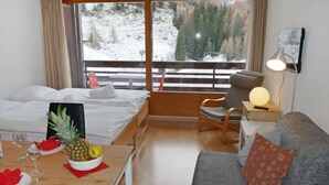 A hotel room with a bed, a dining table with fruit, a sofa, and a view of a snowy landscape through large windows.