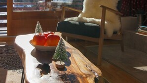 A wooden table with a bowl of apples and two small Christmas trees, a fluffy white chair, and a view of a snowy landscape through a window.
