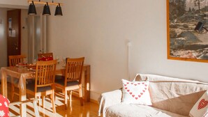 A dining area with wooden chairs and a table, a white sofa with heart-patterned pillows, and a painting of a mountainous landscape on the wall.