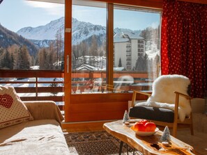 A cozy room with a large window offering a view of snow-covered mountains and a building. There's a plush chair and a sofa with a patterned pillow. - Rosablanche A21 by Interhome (Siviez-Nendaz)