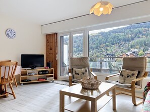 A modern living room with a television, wooden furniture, and a large window with a view of a forested hillside.