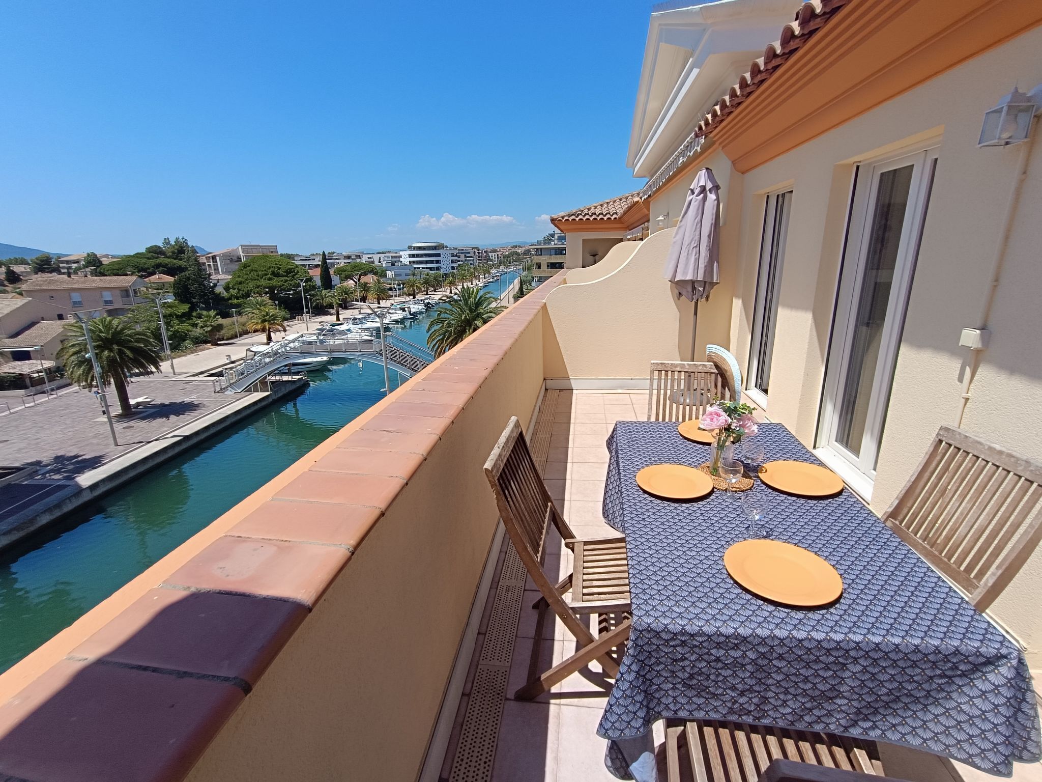 Un balcón con una mesa preparada para dos personas, con vista a un puerto deportivo con barcos y un cielo despejado.