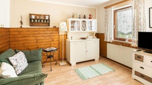 Ein gemütliches Zimmer mit einem grünen Sofa, einem kleinen Tisch, einer Kommode mit Dekoration und einem Fenster mit Vorhängen.