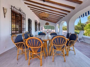 A covered outdoor dining area with wicker chairs and a glass table, featuring a wooden ceiling and arches.